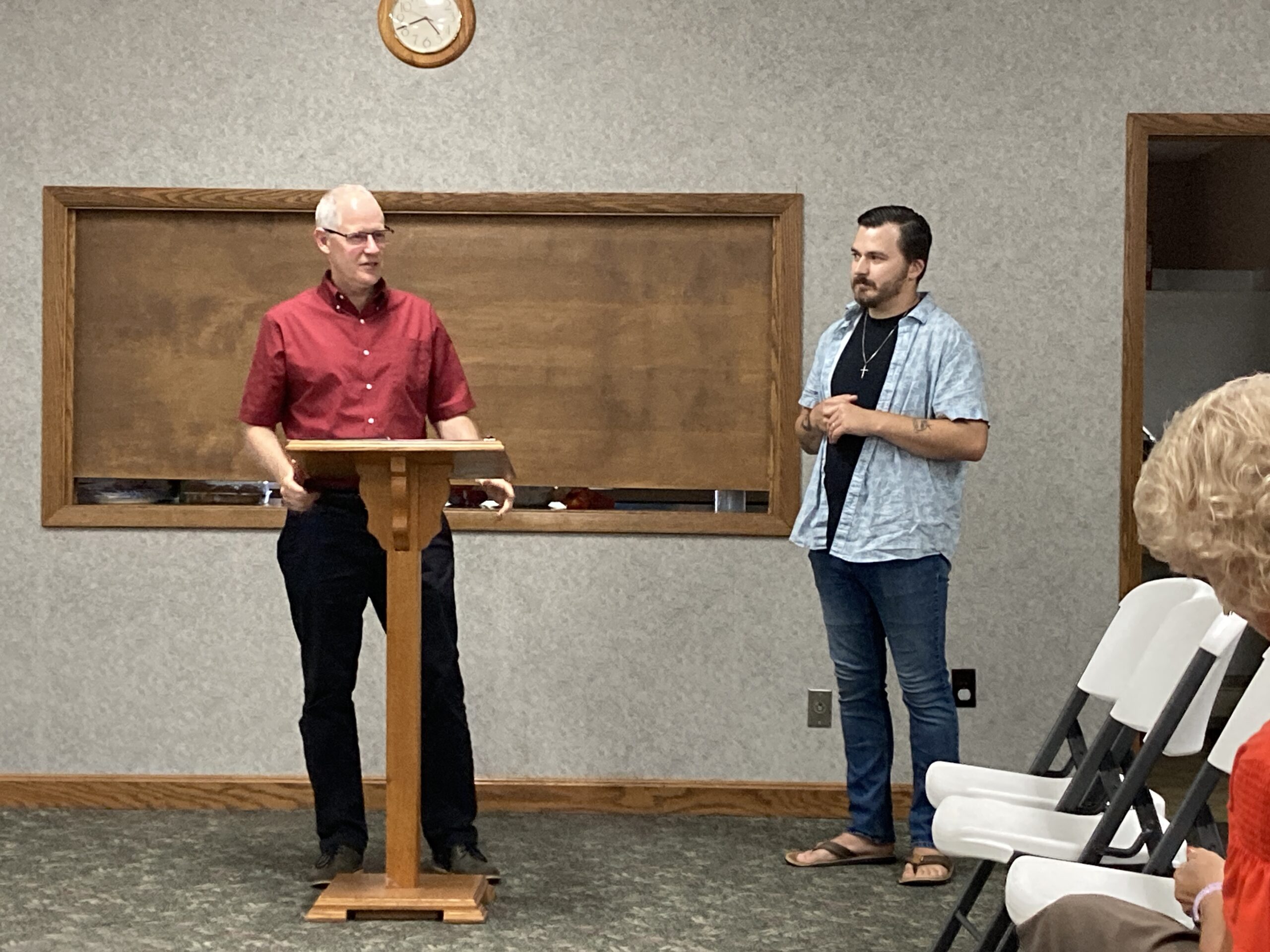Two members of New Life Fellowship at the front of the room during a gathering, one speaking from the podium.