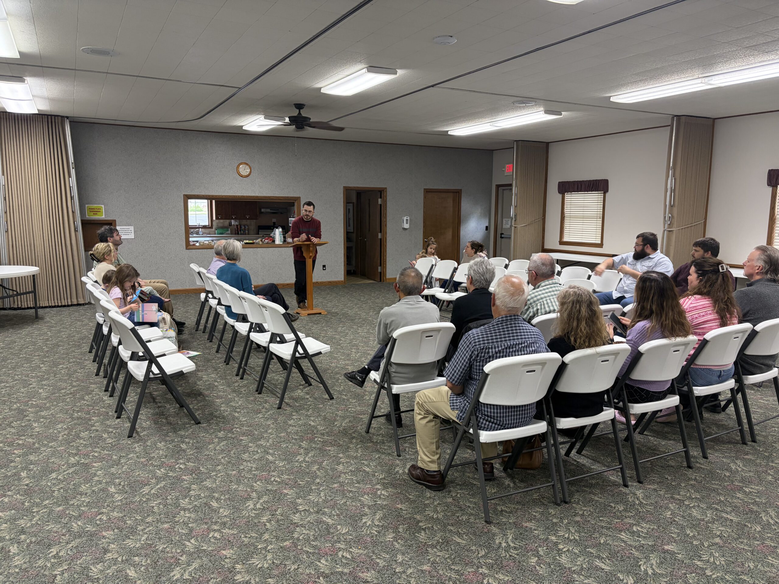 People gathered on folding chairs in the fellowship hall as a speaker leads from a podium at the front.
