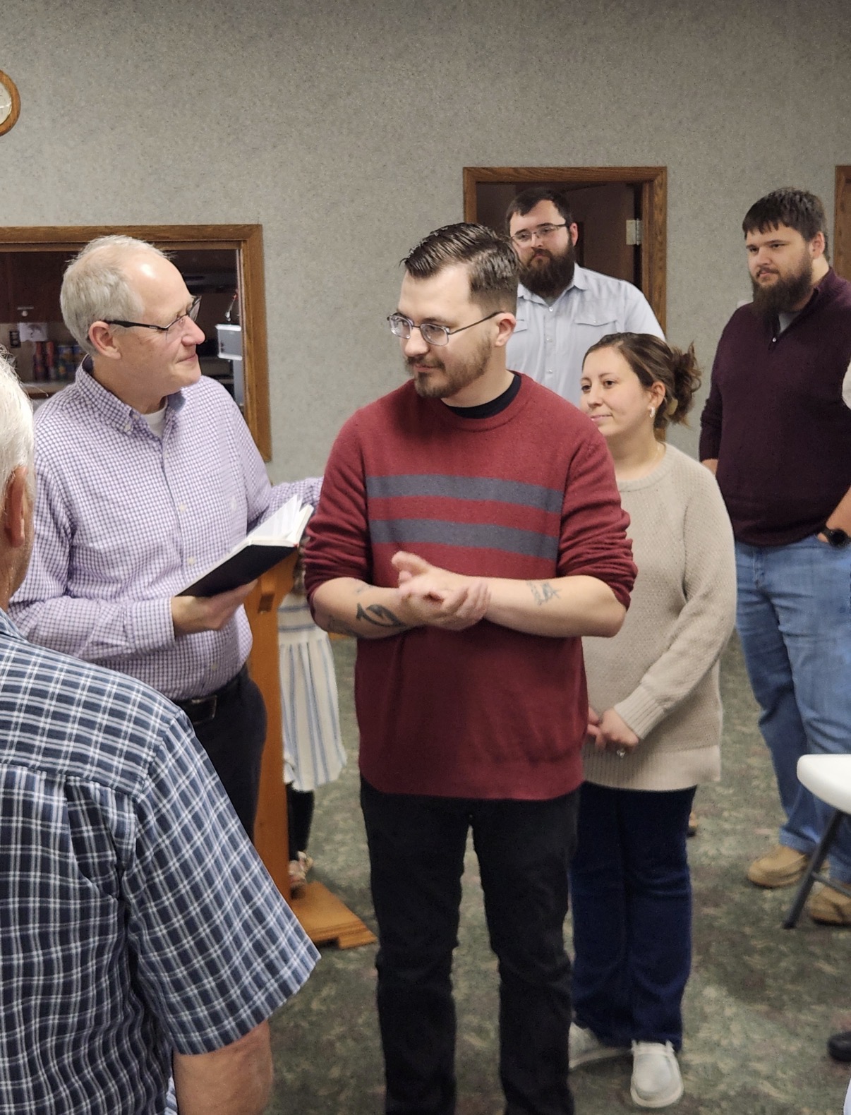Members of New Life Fellowship standing together around Pastor Noah Raines while an older man reads aloud from an open book.